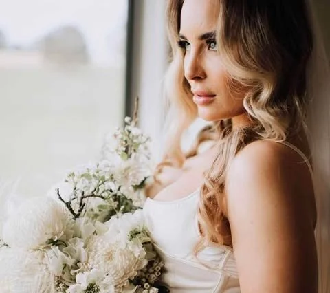 Woman with long, wavy hair holding a bouquet of white flowers, looking out a window.