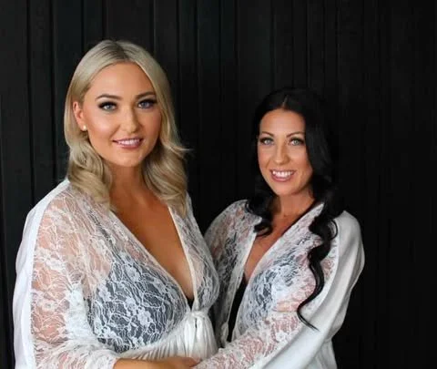 Two women wearing white lace robes standing side by side in front of a black wooden background.