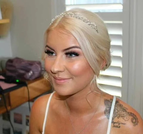 A young woman with platinum blonde hair styled in an elegant updo, wearing a jeweled headband, makeup, and a white spaghetti strap top, standing indoors near a window with shutters.