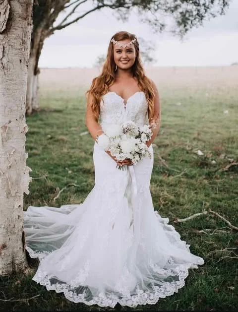 A woman in a white wedding dress holding a bouquet, standing outdoors near a tree with a grassy field in the background.