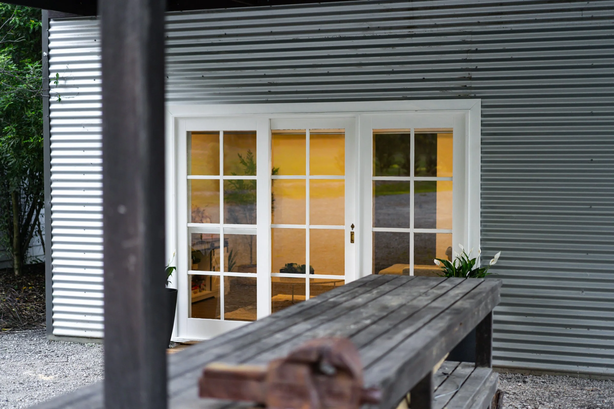 View of a house with a glass door and yellow interior, seen through an outdoor setting with a corrugated metal siding, a wooden table, and some plants.