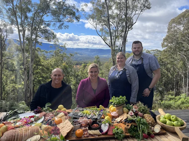 Family enjoying an outdoor meal with a variety of fruits, vegetables, and cheeses on a wooden table in a forested area with mountains in the background.