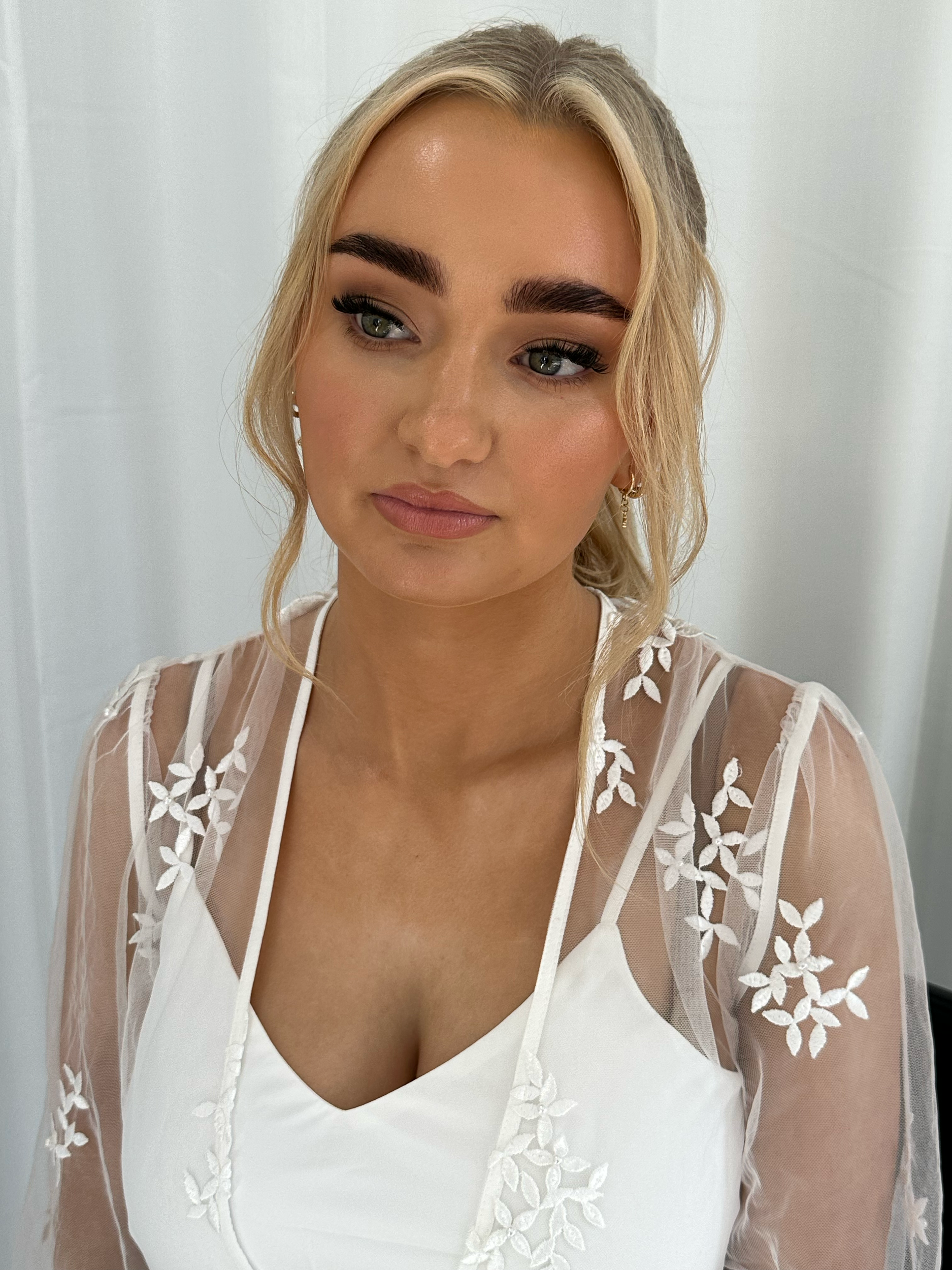 Close-up of a woman with blonde hair, wearing a white dress with a sheer, embroidered overlay with floral patterns, and earrings, standing against a plain background.