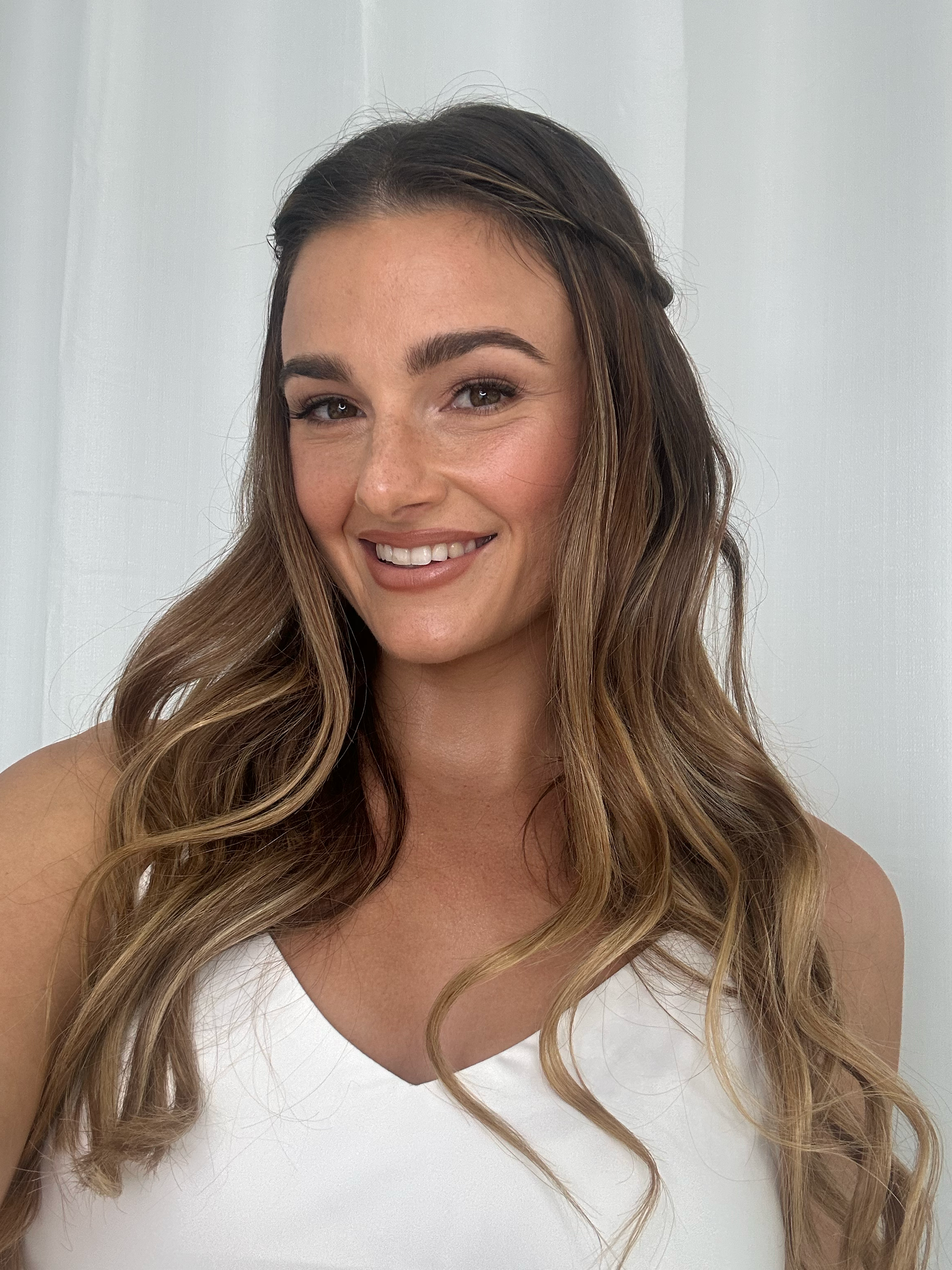 Close-up of a woman with long, wavy, light brown hair, smiling, wearing a white top, standing against a plain light background.