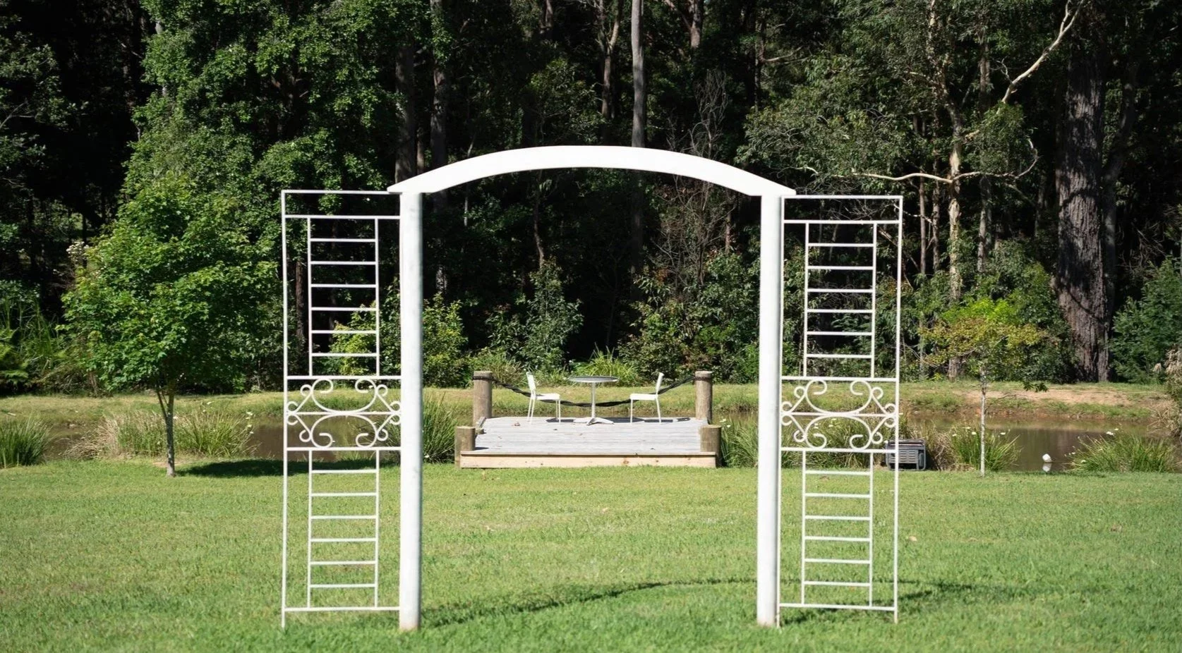 White garden archway with lattice sides leading to a wooden deck with a table and two chairs, set in a green grassy area near a pond and surrounded by trees and bushes.