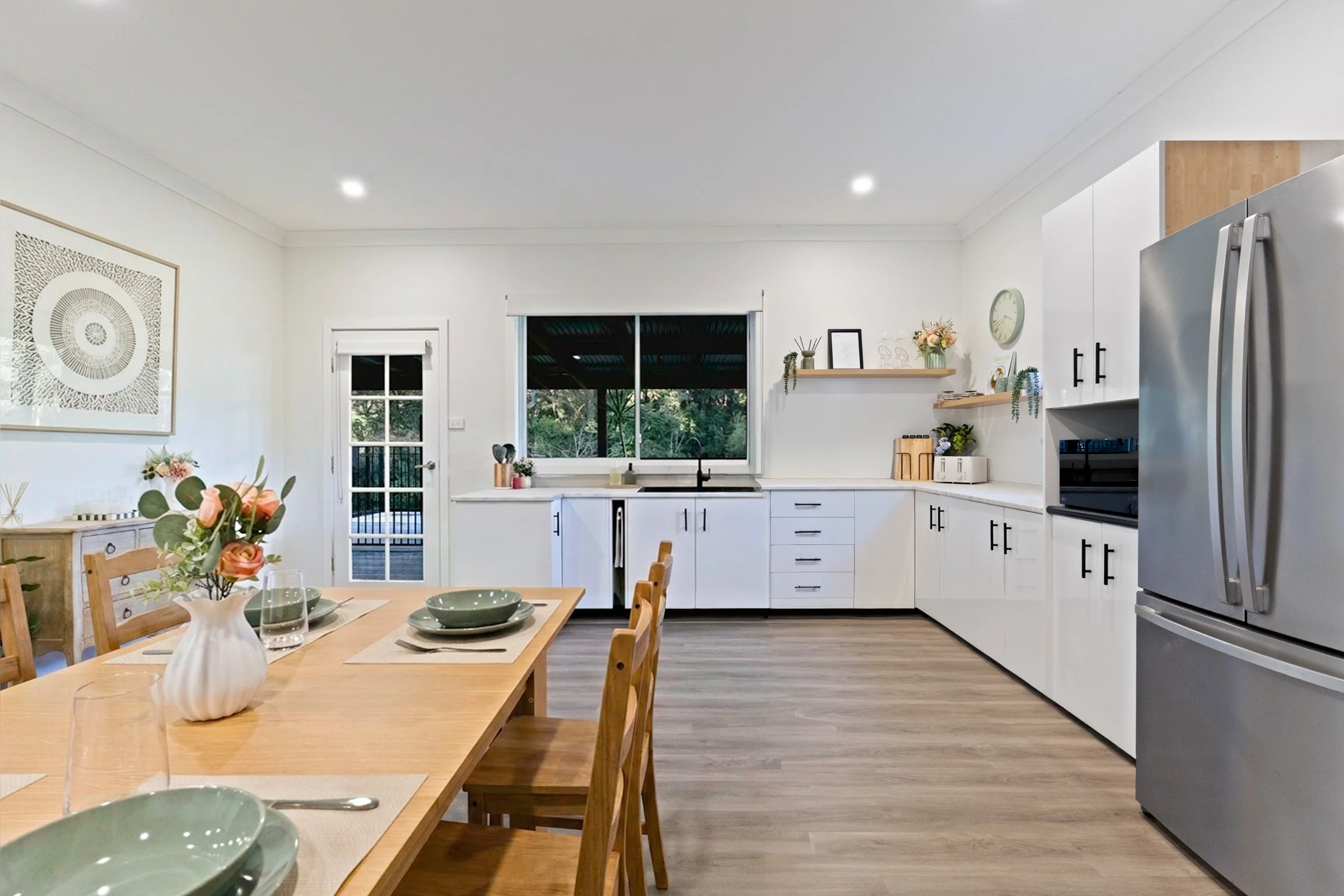 A bright, modern kitchen with white cabinets, a stainless steel refrigerator, a window showing greenery outside, and a dining table with chairs and a floral centerpiece.