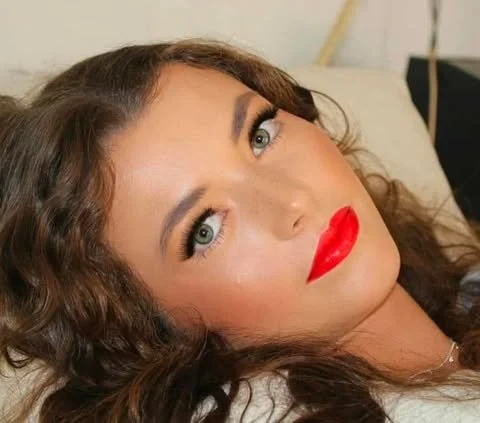 Close-up of a woman with wavy brown hair, bright red lipstick, and eye makeup, lying on a bed or couch.