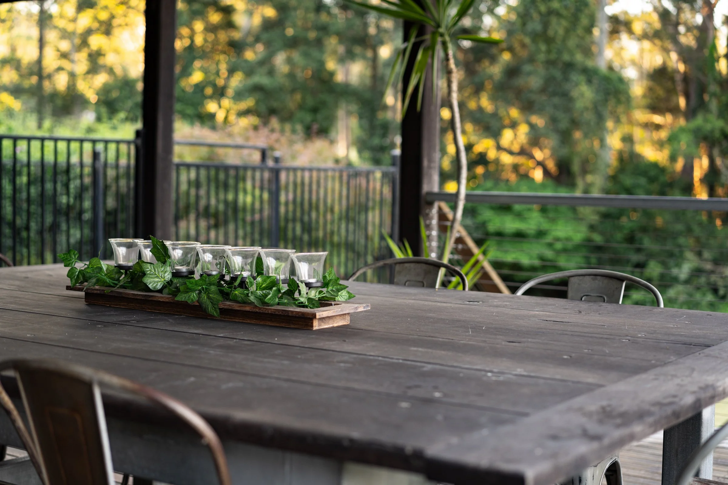 An outdoor wooden table with a tray of small candle jars surrounded by green ivy, set on a porch or deck with a black metal railing, overlooking a forested area with sunlight filtering through the trees.