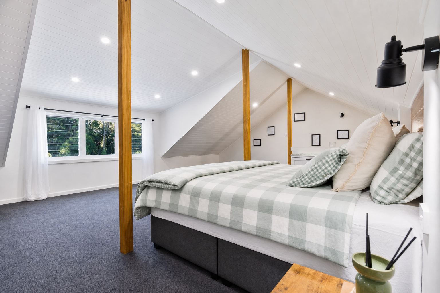A bedroom with a large bed featuring checkered bedspread and pillows, situated near windows with white curtains, and a gallery wall with framed pictures on the white wall, in a room with a vaulted ceiling and wooden support beams.