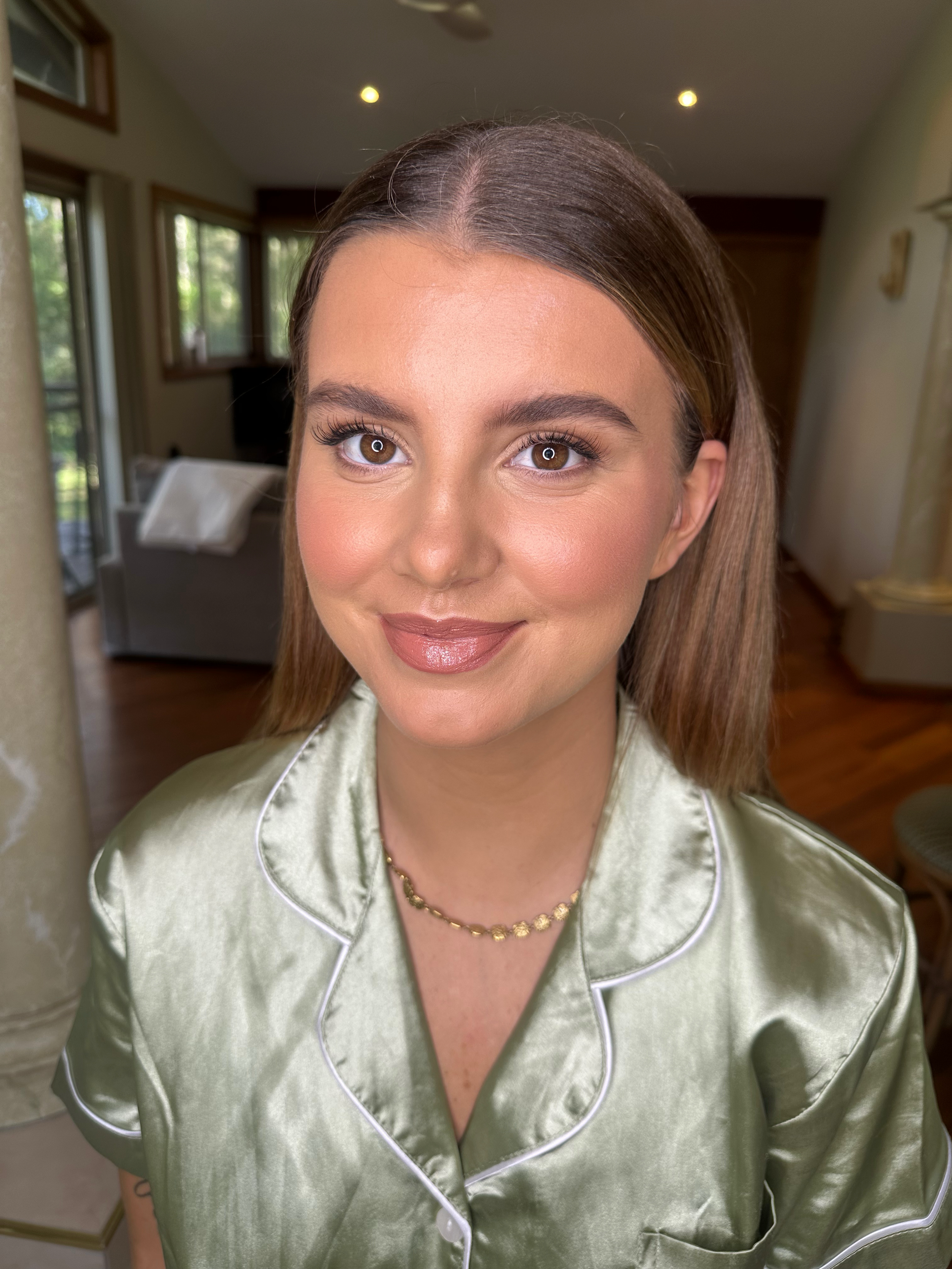 Close-up of a woman's face with brown hair in a bedroom, wearing satin pajamas and a gold necklace.