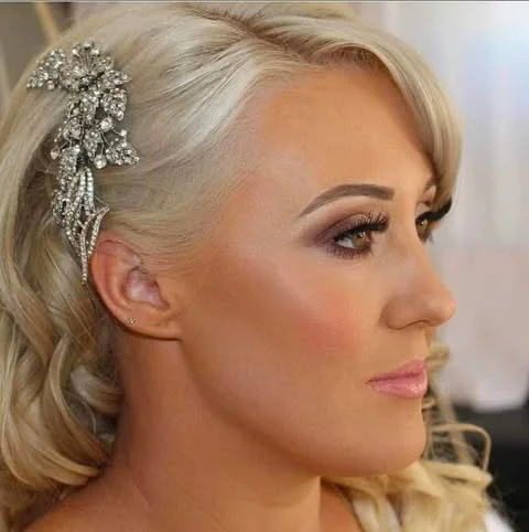 Close-up of a woman with blonde hair styled in loose waves, wearing a decorative hairpiece with silver embellishments, and makeup with neutral eyeshadow, defined brows, and pink lipstick.