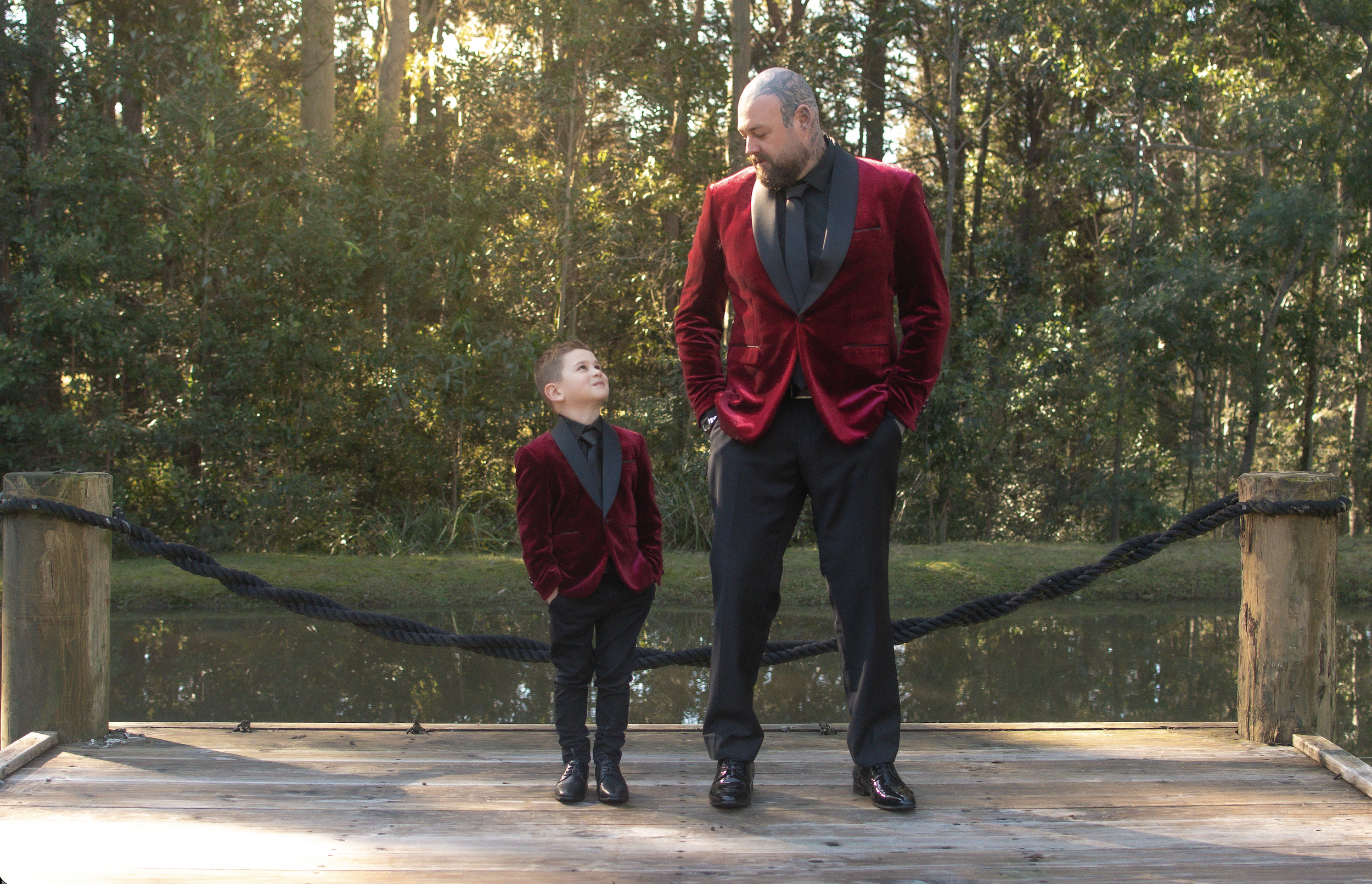 A man in a red velvet blazer and a young boy in a matching red velvet blazer walk together on a wooden bridge in a wooded area during daytime. The man looks down at the boy, who looks up at him, with trees in the background and sunlight filtering thr