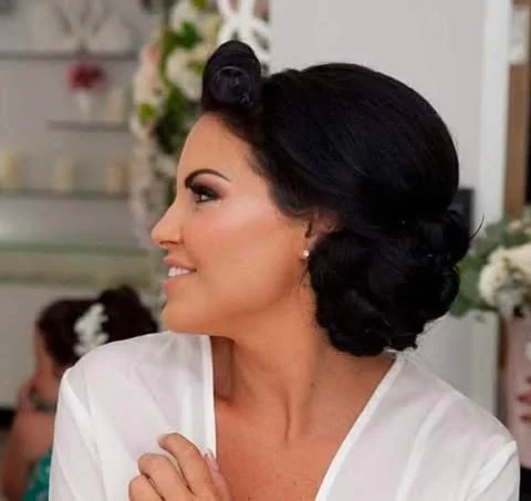 Side profile of a woman with dark hair styled in vintage waves, wearing a white robe, smiling and looking to the left, with a blurred background of people and floral decorations.