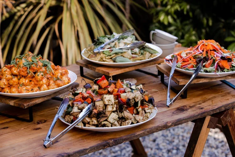 Outdoor table with four plates of various salads and dishes, including chickpea salad, vegetable medley, mixed grilled vegetables, and a leafy green salad, set against greenery background.
