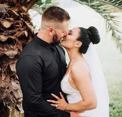 A bride and groom sharing a kiss outdoors near a palm tree, with the bride in a wedding dress and veil, and the groom in a black suit.