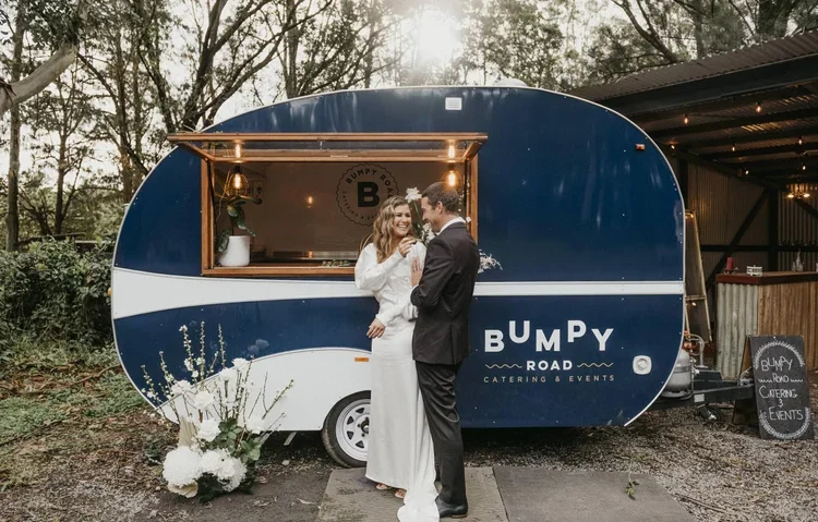 A couple in wedding attire stands in front of a blue mobile food trailer named Bumpy Road Catering & Events, decorated with flowers and string lights, outdoors near trees and a rustic building at sunset.