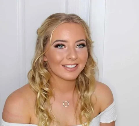 Young woman with blonde hair, blue eyes, and a soft smile, wearing a white off-the-shoulder top and a circular necklace, standing in front of a white background.