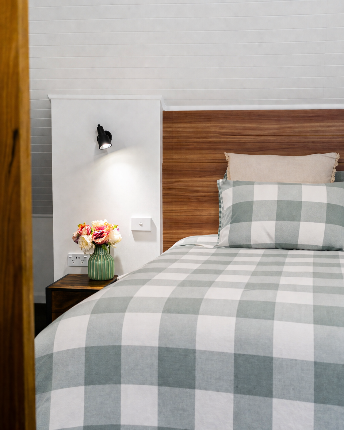 Close-up of a neatly made bed with a checkered gray and white duvet, beige pillow, and wooden headboard in a modern bedroom. A nightstand with a green striped vase holding pink and white flowers, a black wall-mounted reading light, and a white wall w
