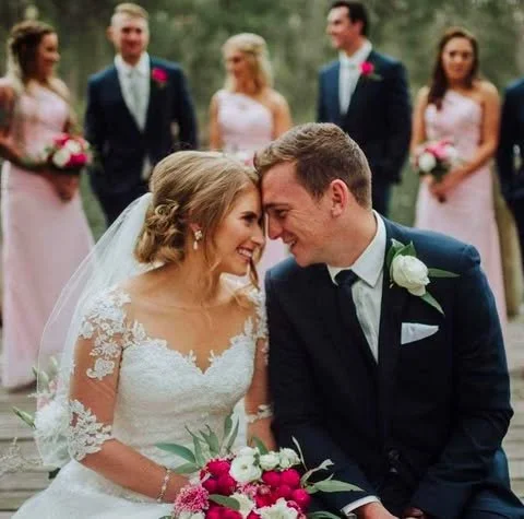 Bride and groom smiling and leaning their foreheads together at their wedding, with bridesmaids and groomsmen in the background.