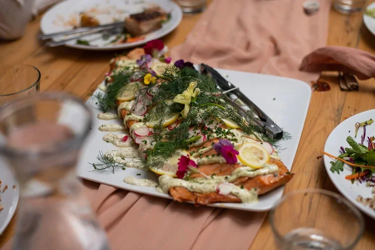 A whole cooked fish garnished with herbs, lemon slices, radish slices, edible flowers, and creamy sauce, served on a white platter on a wooden table.