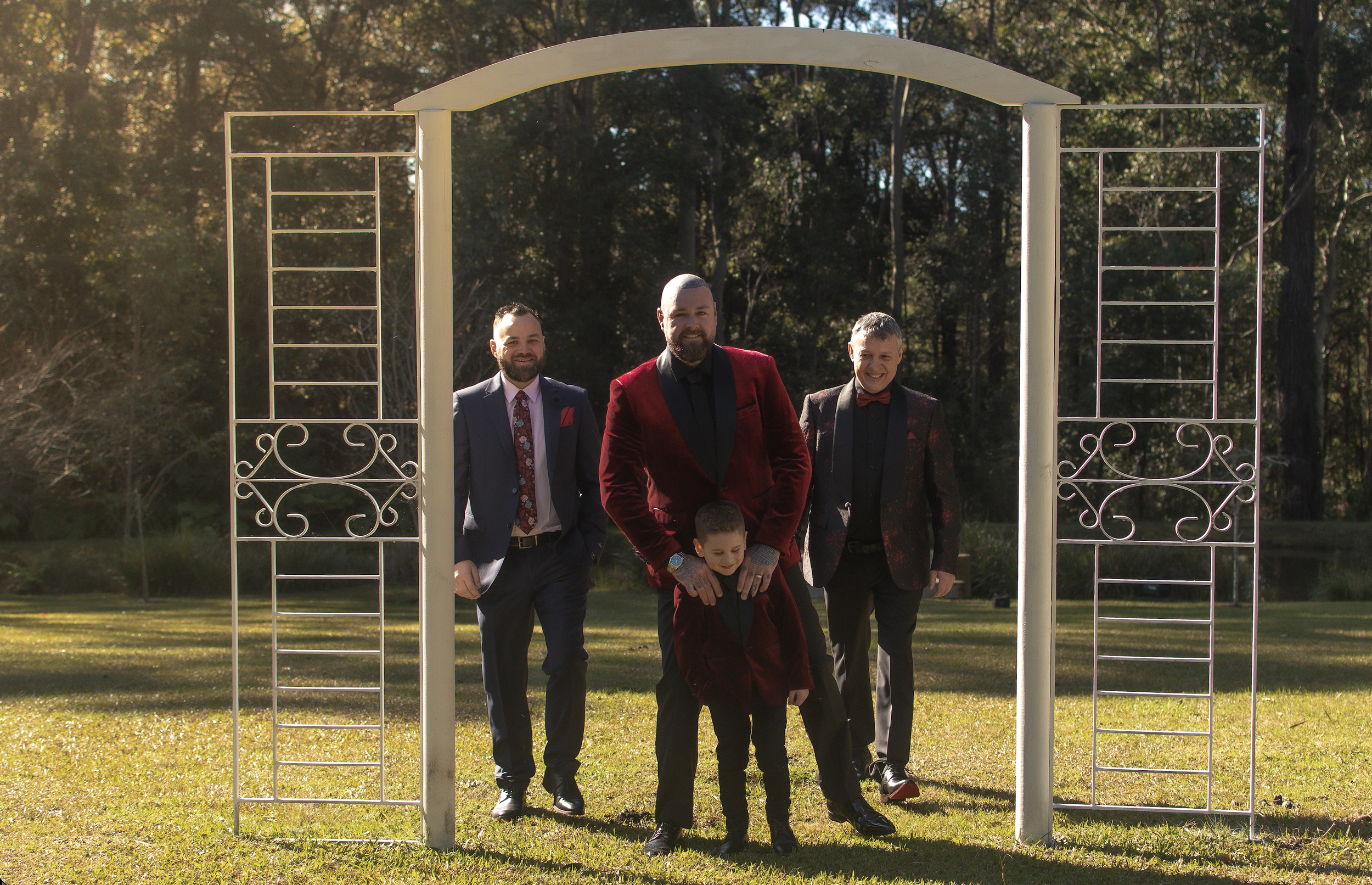 Three men and a boy walk through a decorative garden arch on a grassy area, with trees in the background and sunlight filtering through.