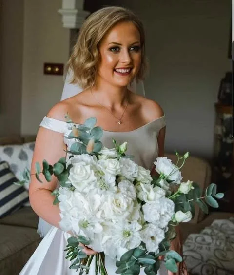 A woman in a wedding dress holding a bouquet of white flowers and eucalyptus leaves, smiling indoors