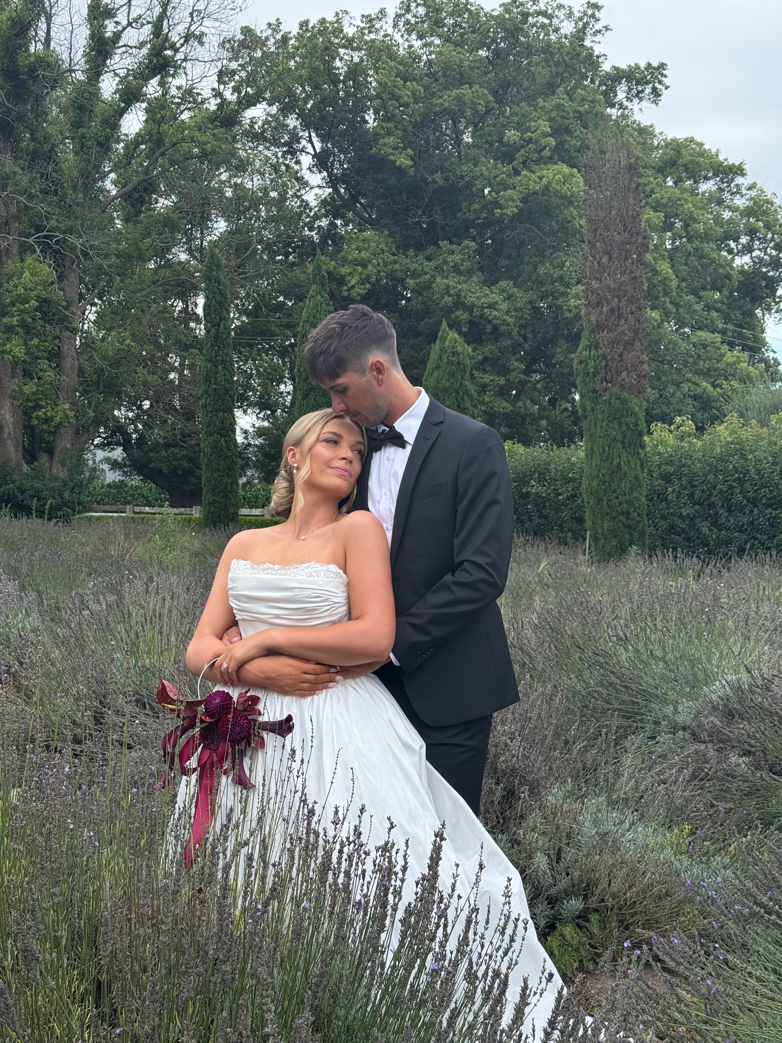 A bride and groom embrace in a garden with tall purple flowers and green trees in the background during a wedding photo shoot.