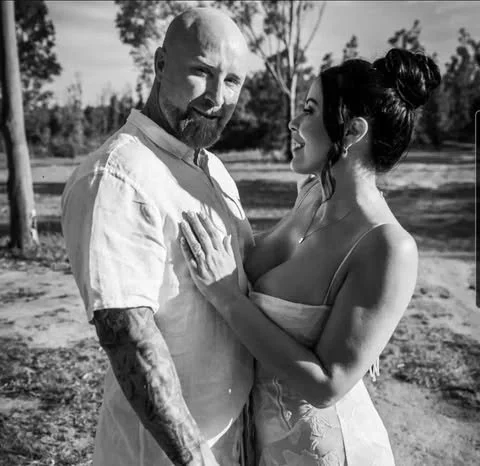 A man and woman standing outdoors, looking at each other affectionately. The man has a beard, tattoos, and is wearing a short-sleeve shirt. The woman has her hair in a bun and is wearing a strapless top or dress.