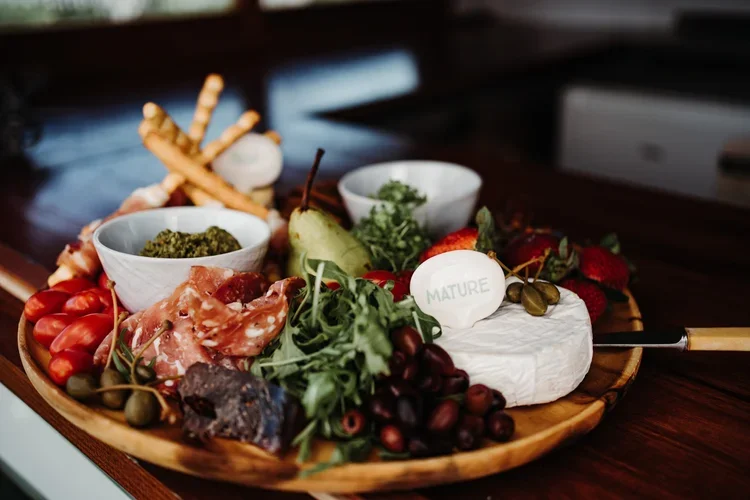 A wooden platter with cheese, grapes, cherry tomatoes, prosciutto, breadsticks, and condiments.