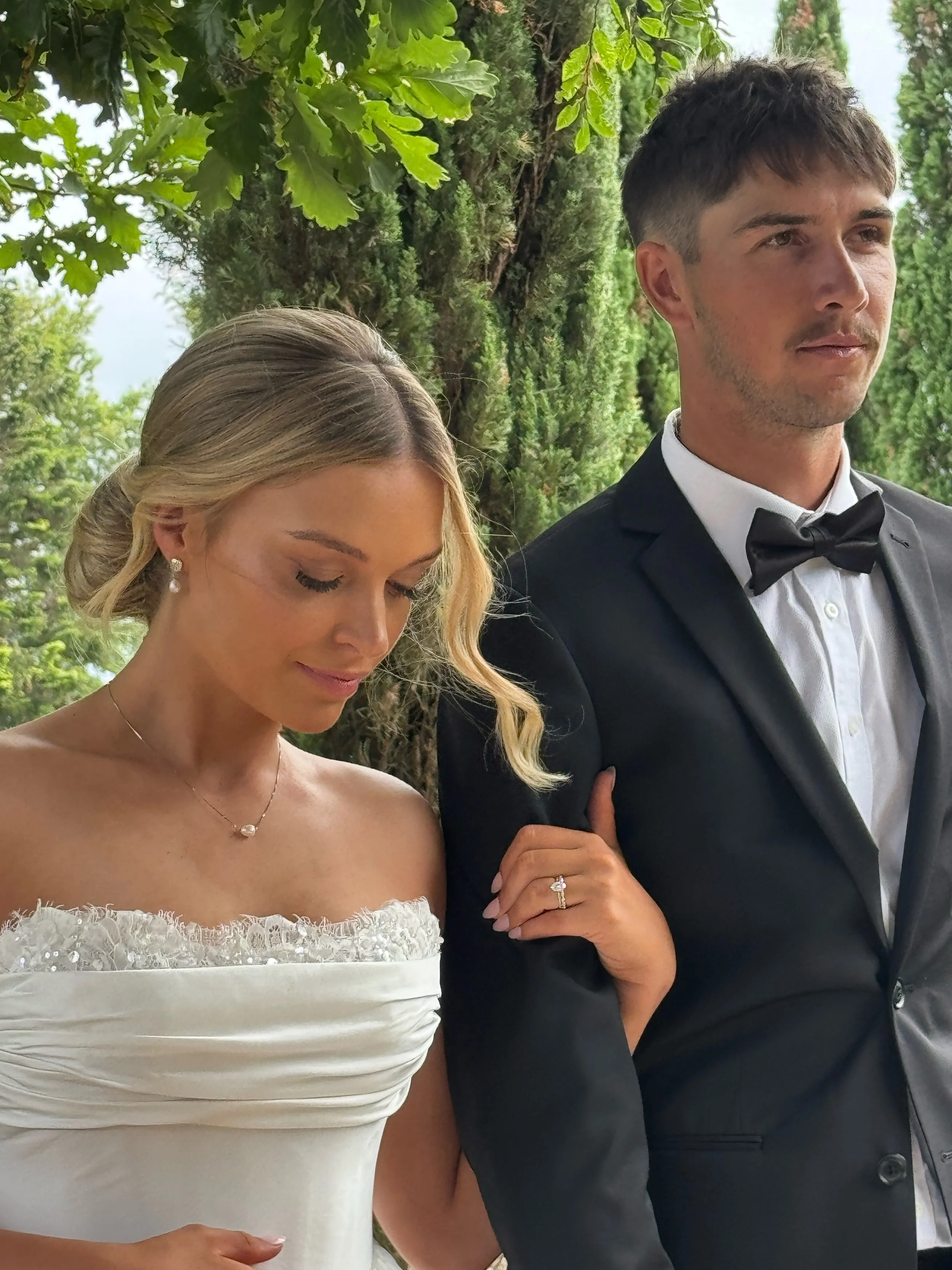 A close-up of a bride and groom holding hands, standing outdoors amidst lush green trees. The bride has blonde hair styled in loose curls, wearing elegant pearl earrings and a necklace, and a strapless white wedding dress with lace detailing. The gro