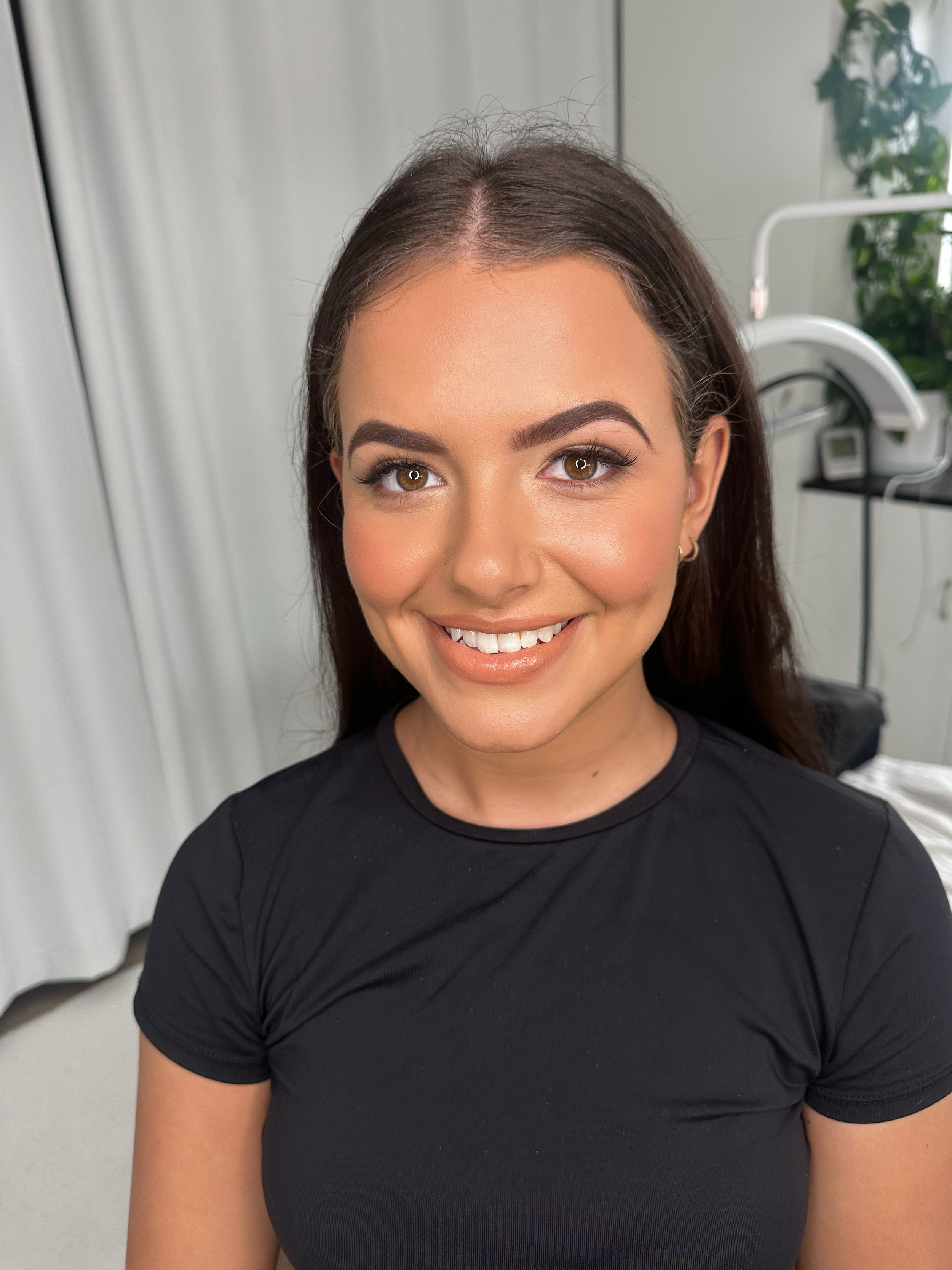 Close-up of a young woman with long dark hair, smiling, wearing a black t-shirt, in a room with medical equipment and a plant in the background.