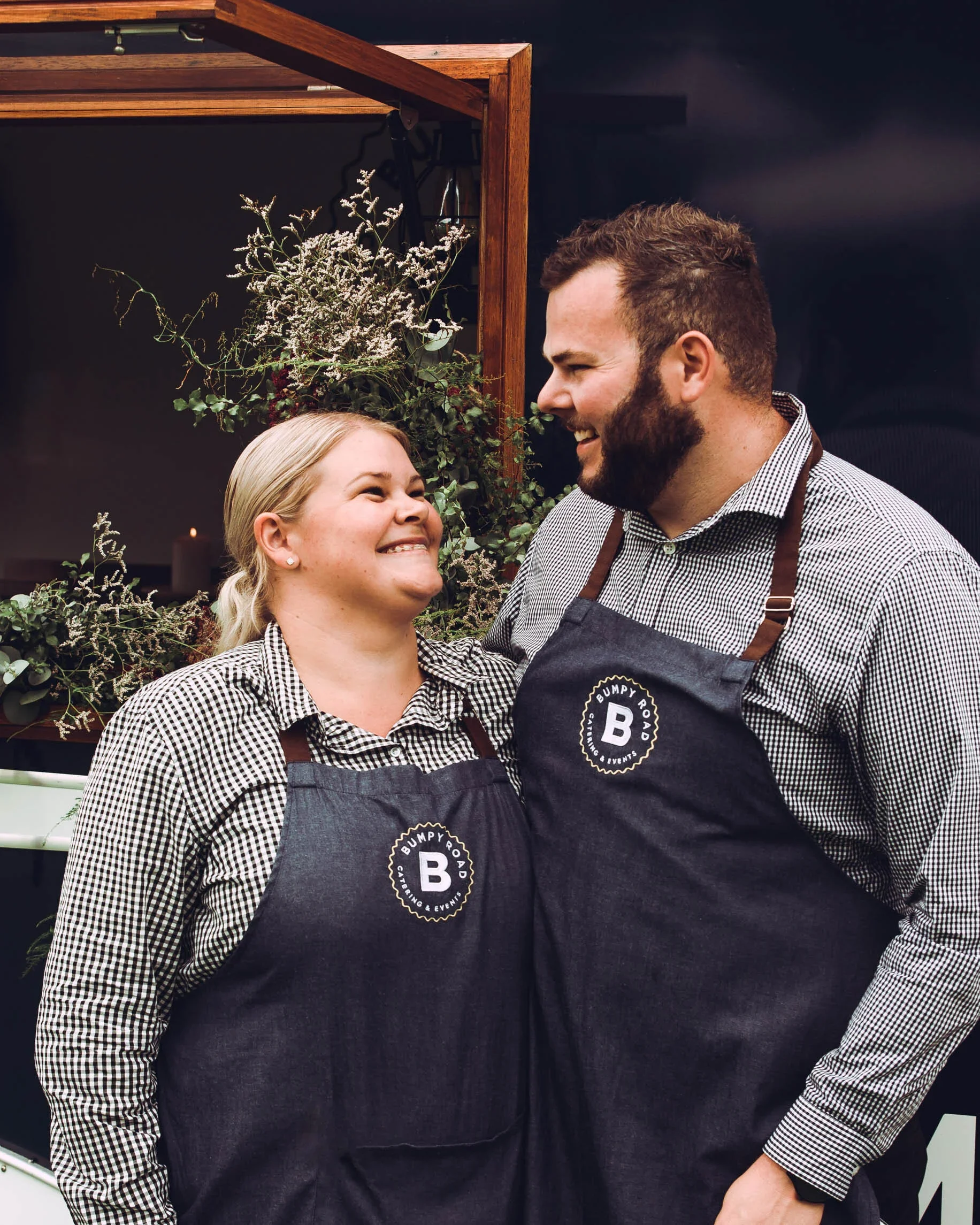 Smiling man and woman wearing black aprons with a logo, standing closely and looking at each other, with a floral arrangement in the background.
