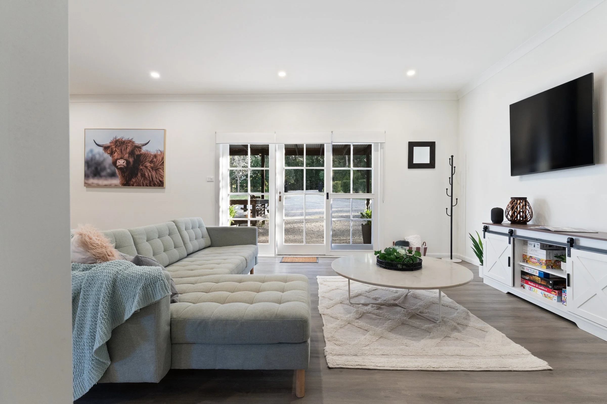 A living room with a large beige tufted sectional sofa, a round coffee table, a white rug, a wall-mounted flat-screen TV, a barn-style white TV stand with books and decor, a coat rack, potted plants, and a window with sliding glass doors leading outs