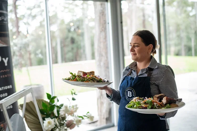 A woman in a checkered shirt and blue apron holding two plates of salad in a restaurant with large windows and trees outside.
