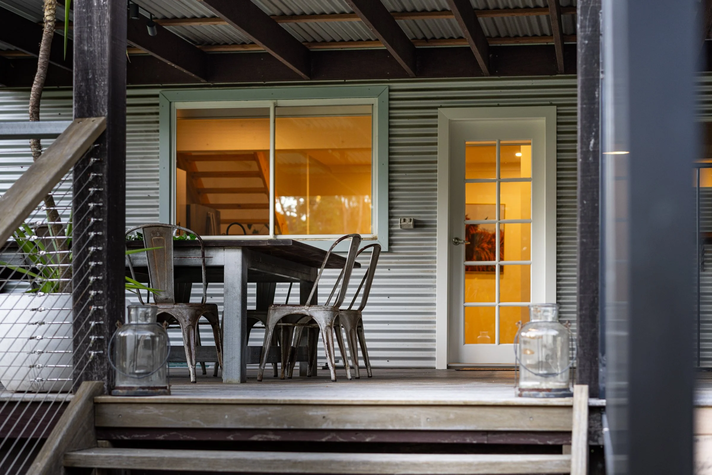 Outdoor patio with metal table, chairs, lanterns, and a building with sliding window and door, illuminated from inside.
