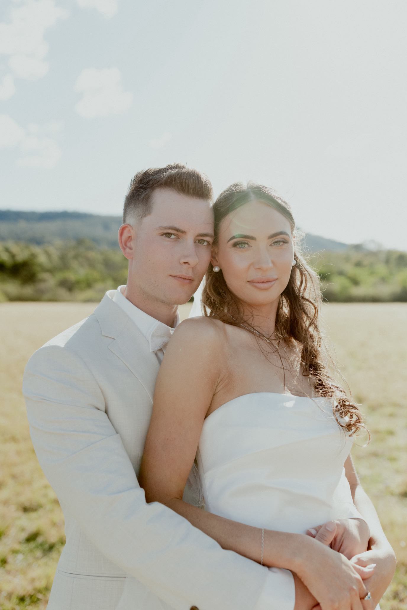 A couple dressed in wedding attire outdoors during daytime. The man is wearing a light-colored suit, and the woman is in a strapless white wedding dress. They are standing close, with the man's arm around the woman's waist, both looking into the came