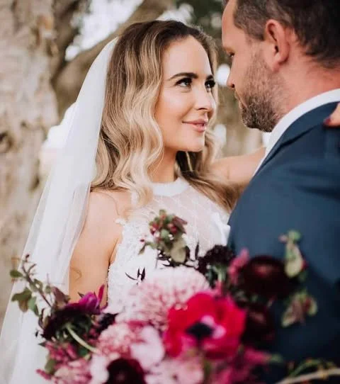 Bride and groom facing each other outdoors, surrounded by trees, with the bride holding a bouquet of pink, purple, and white flowers.