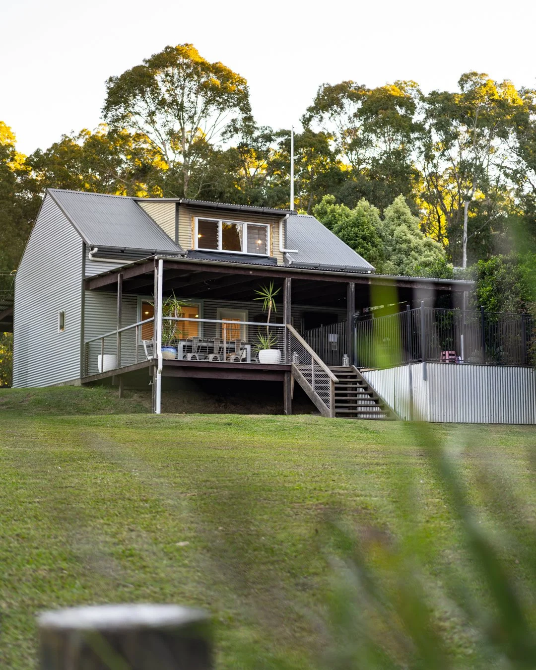 A modern house with a metal roof and a large deck, surrounded by trees, with a grassy yard in the foreground.