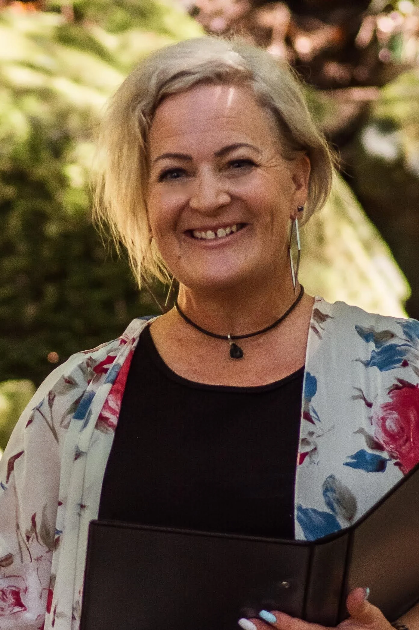 A smiling middle-aged woman with short blonde hair, wearing a black shirt, a floral jacket, and a choker necklace, holding a closed black laptop in front of her, with a background of blurred greenery.