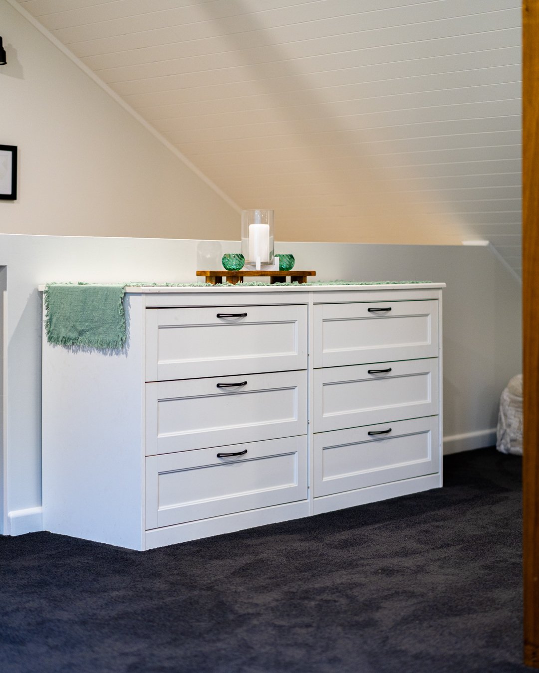 A white dresser with six drawers, black handles, and a green cloth draped over the left side. On top of the dresser, there's a wooden tray with a large candle in a glass holder and two small green glass containers. The room has a sloped ceiling with 