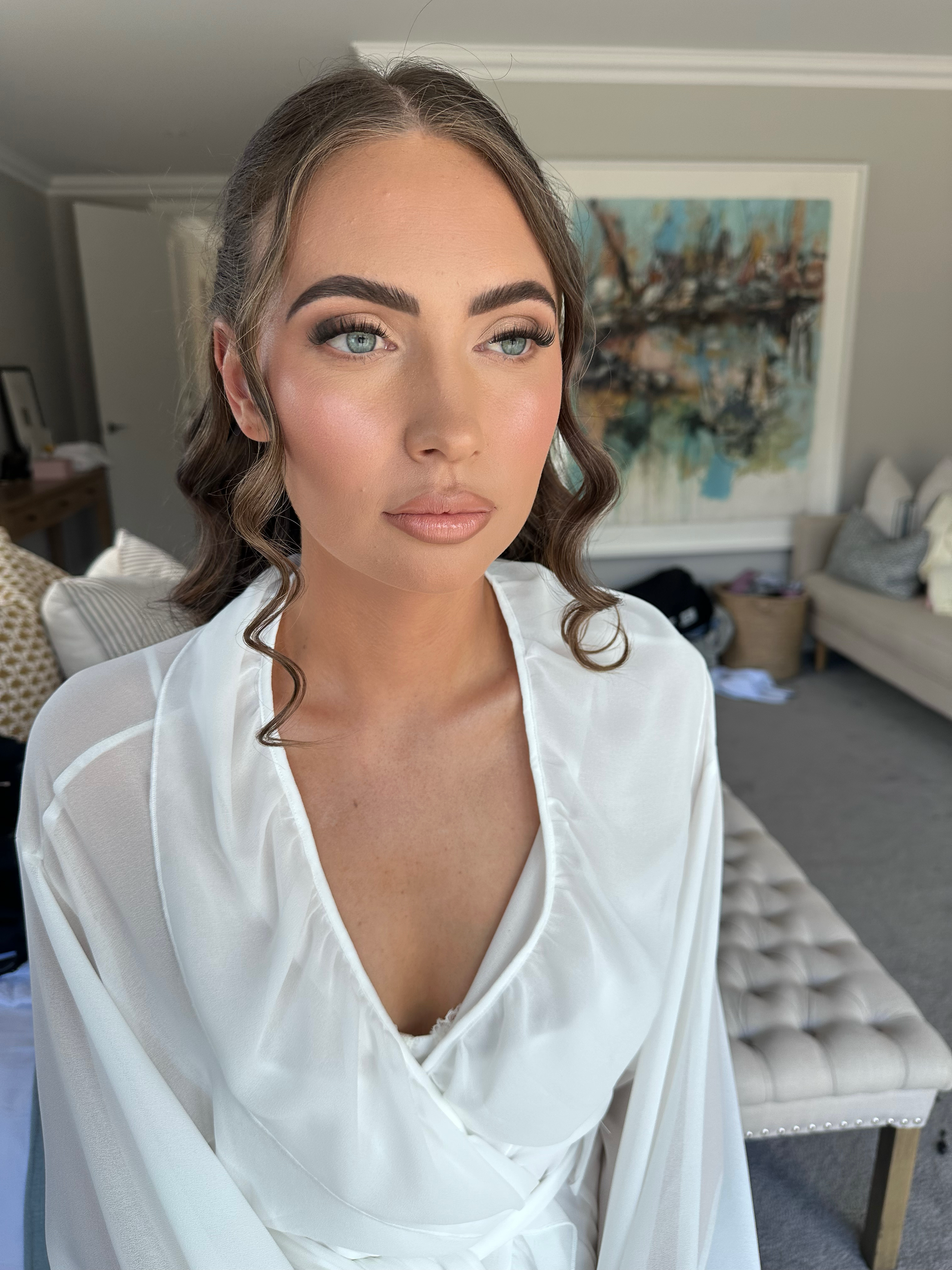 Close-up of a woman with brown hair styled in loose waves, wearing makeup, and dressed in a white robe, sitting in a room with artwork on the wall and furniture in the background.