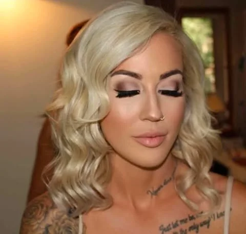 Close-up of a woman with platinum blonde hair styled in loose waves, wearing makeup with dark eyeliner, eyeshadow, and natural lipstick, in an indoor setting with a blurred background.