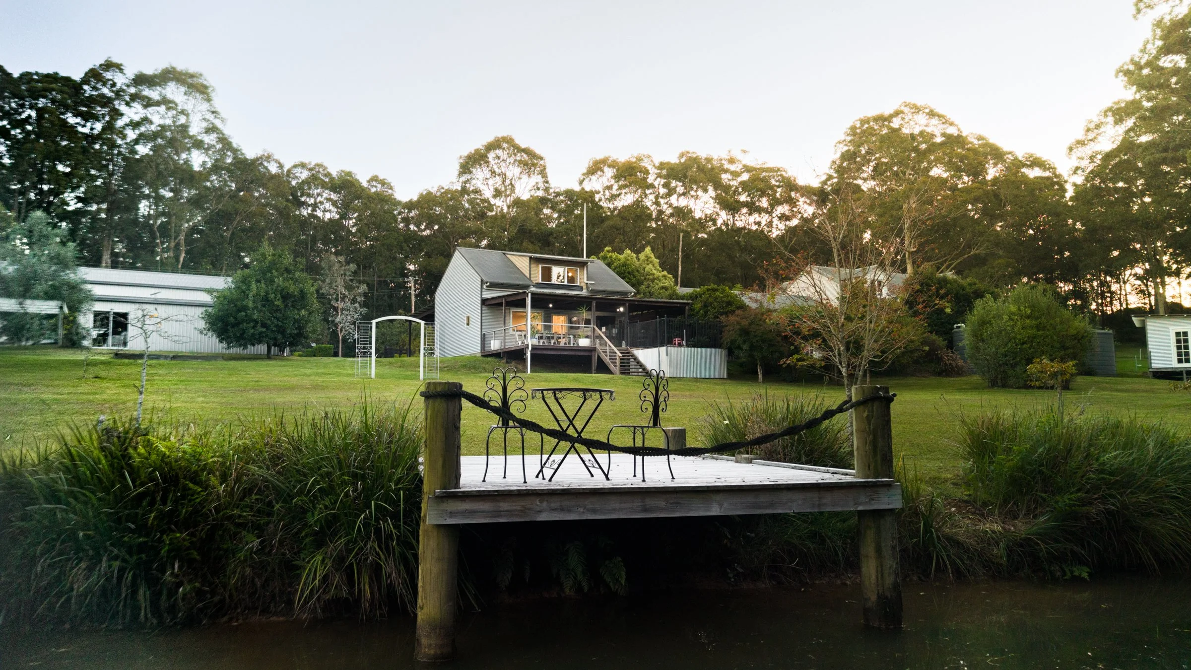 A house with a deck overlooking a grassy yard and trees, with a small pond or stream in the foreground and outdoor furniture on the deck, during sunset or early evening.