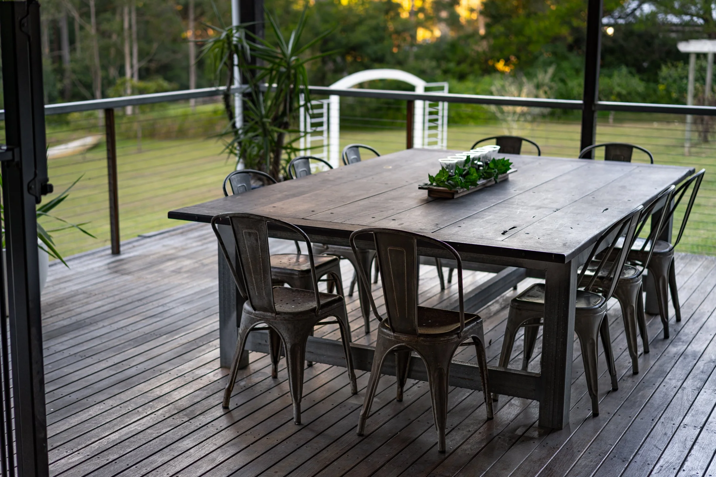 An outdoor dining area with a large wooden table and metal chairs on a deck, surrounded by a railing and greenery in the background.