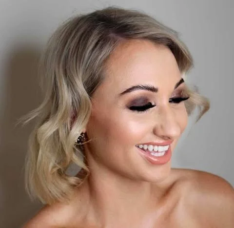 A smiling woman with blonde, wavy hair and makeup, looking down and to the side.