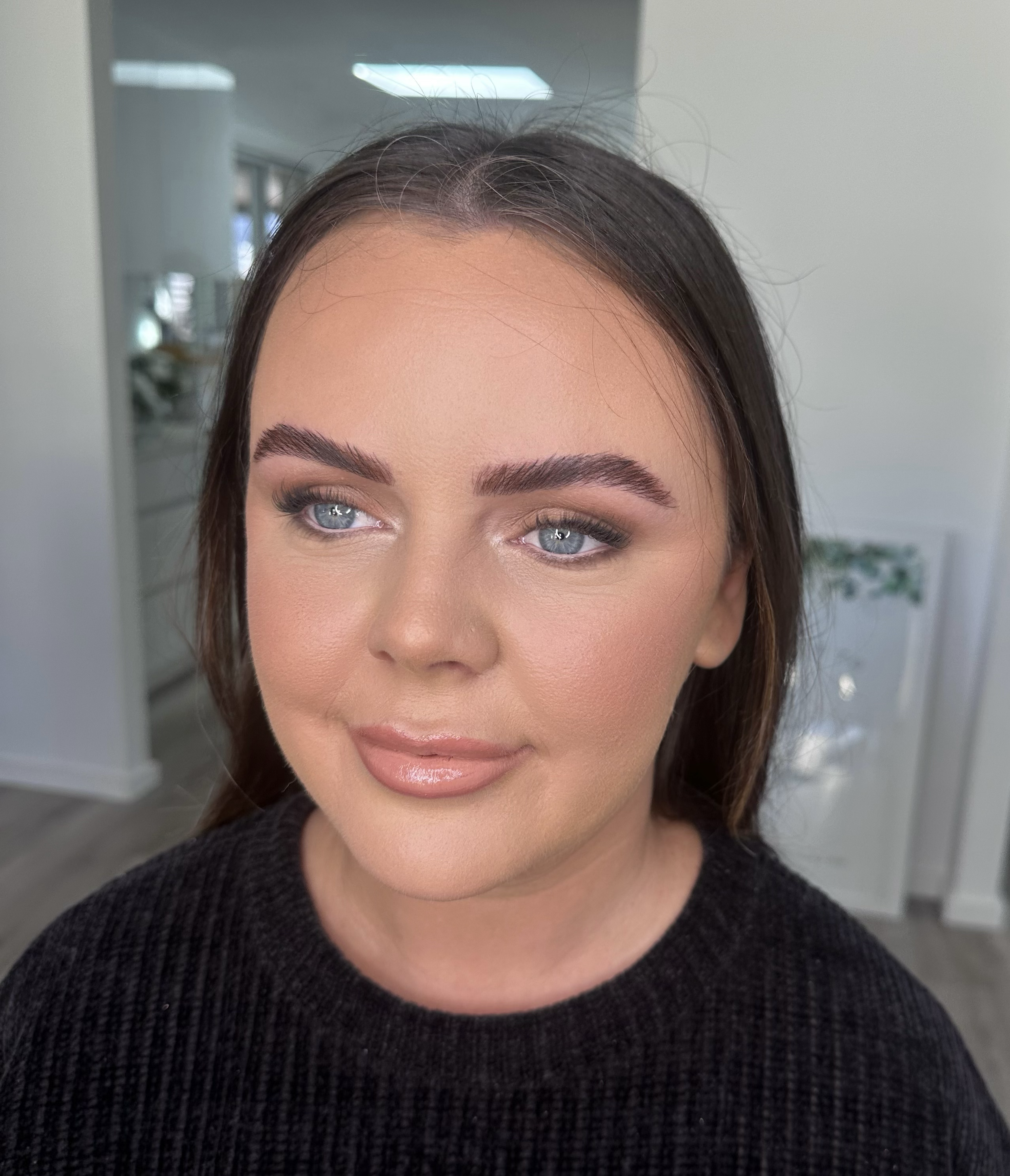 Close-up of a woman's face showing her forehead, eyes, and part of her nose and mouth. She has light skin, blue eyes, and dark brown hair, with makeup including eyeshadow, mascara, and lipstick. She is wearing a black textured top.