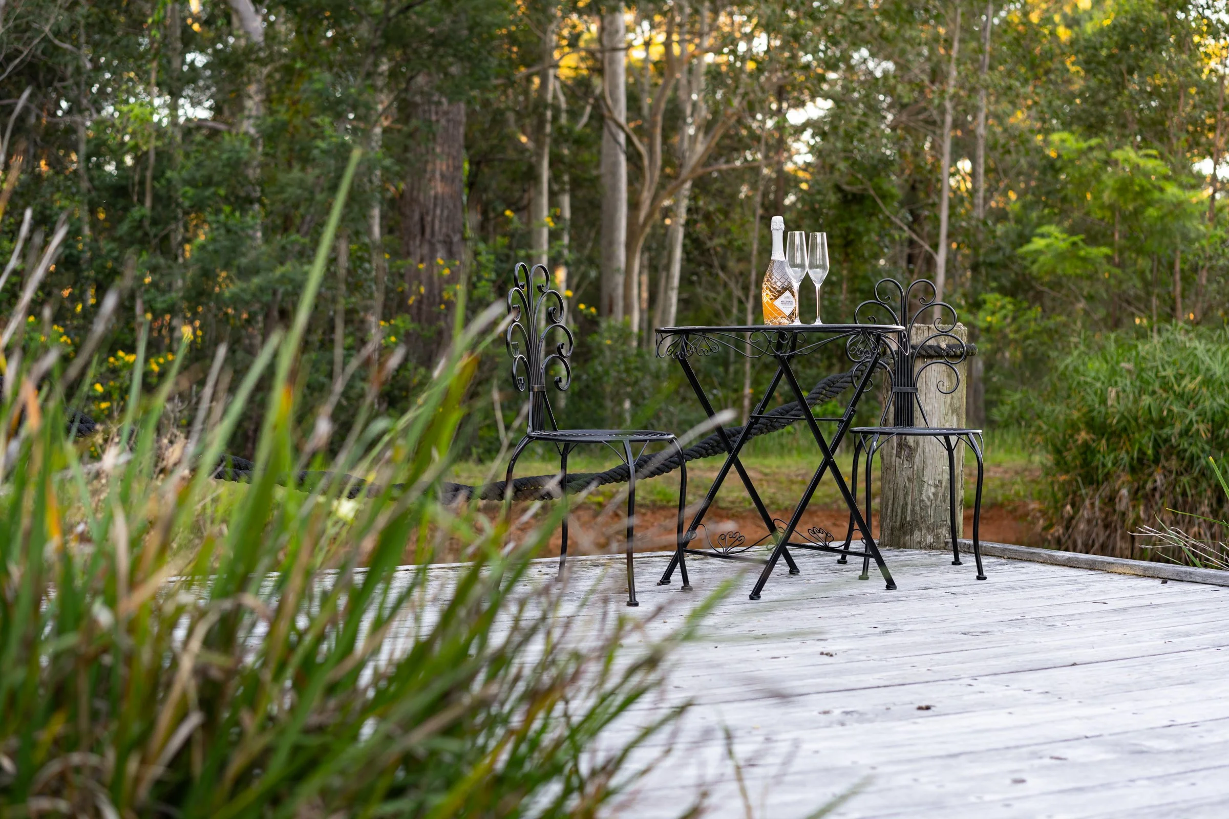 Outdoor scene with a black wrought iron table and chairs on a wooden deck, with a bottle of champagne and two champagne flutes, surrounded by trees in a lush green forest setting.