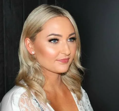 A young woman with blonde hair, wearing makeup, and a white lace top, standing against a dark background.