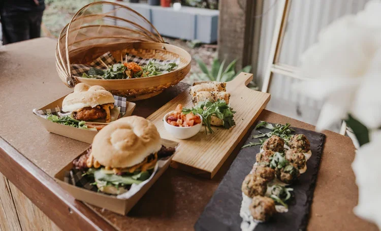 Assorted sandwiches, meatballs on a black platter, and a salad in a wooden and glass bowl on a wooden table.