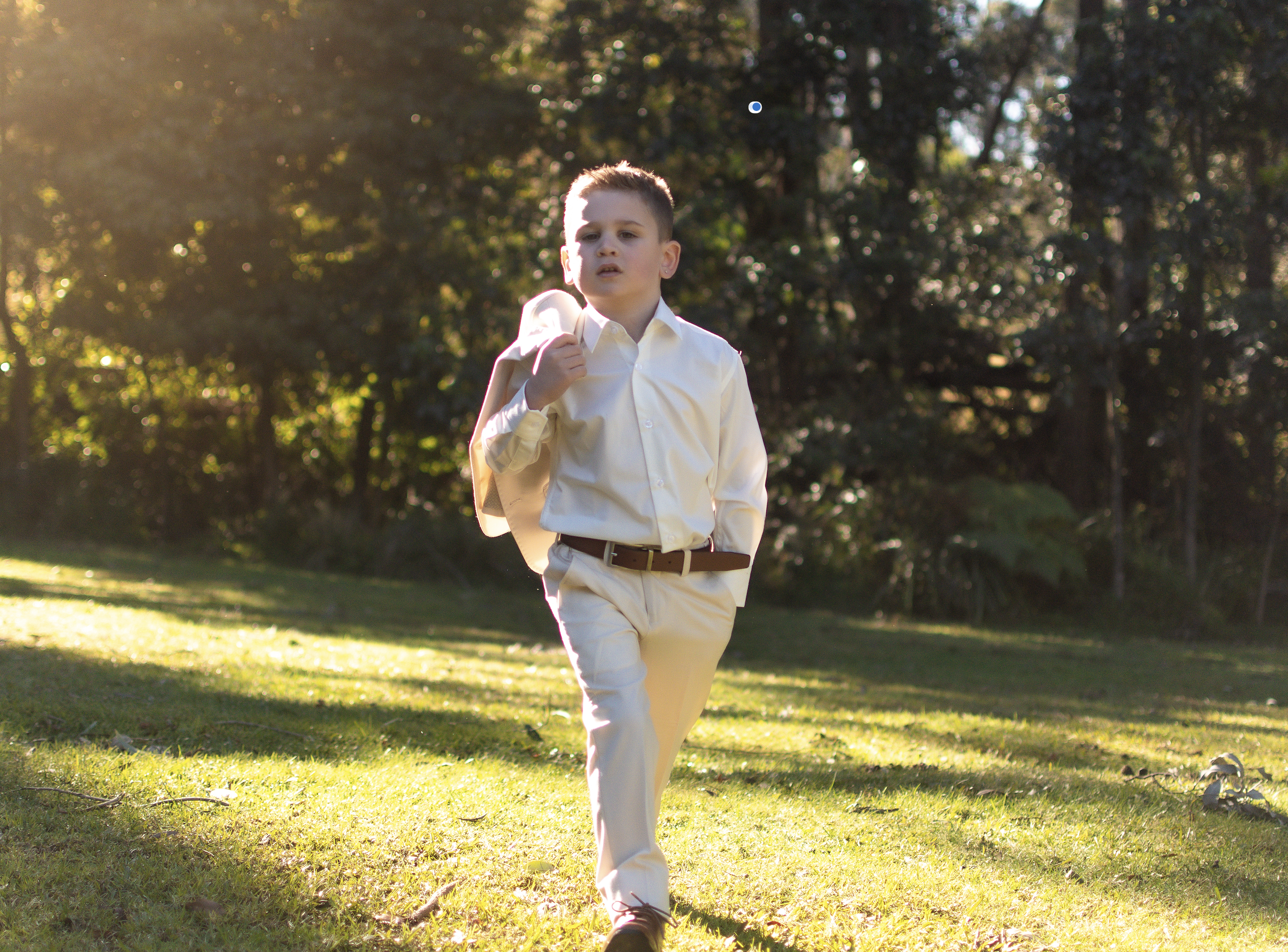 A young boy in a white dress shirt and light-colored pants walking outdoors on a grassy area, carrying a jacket over his shoulder, with sunlight filtering through trees in the background.